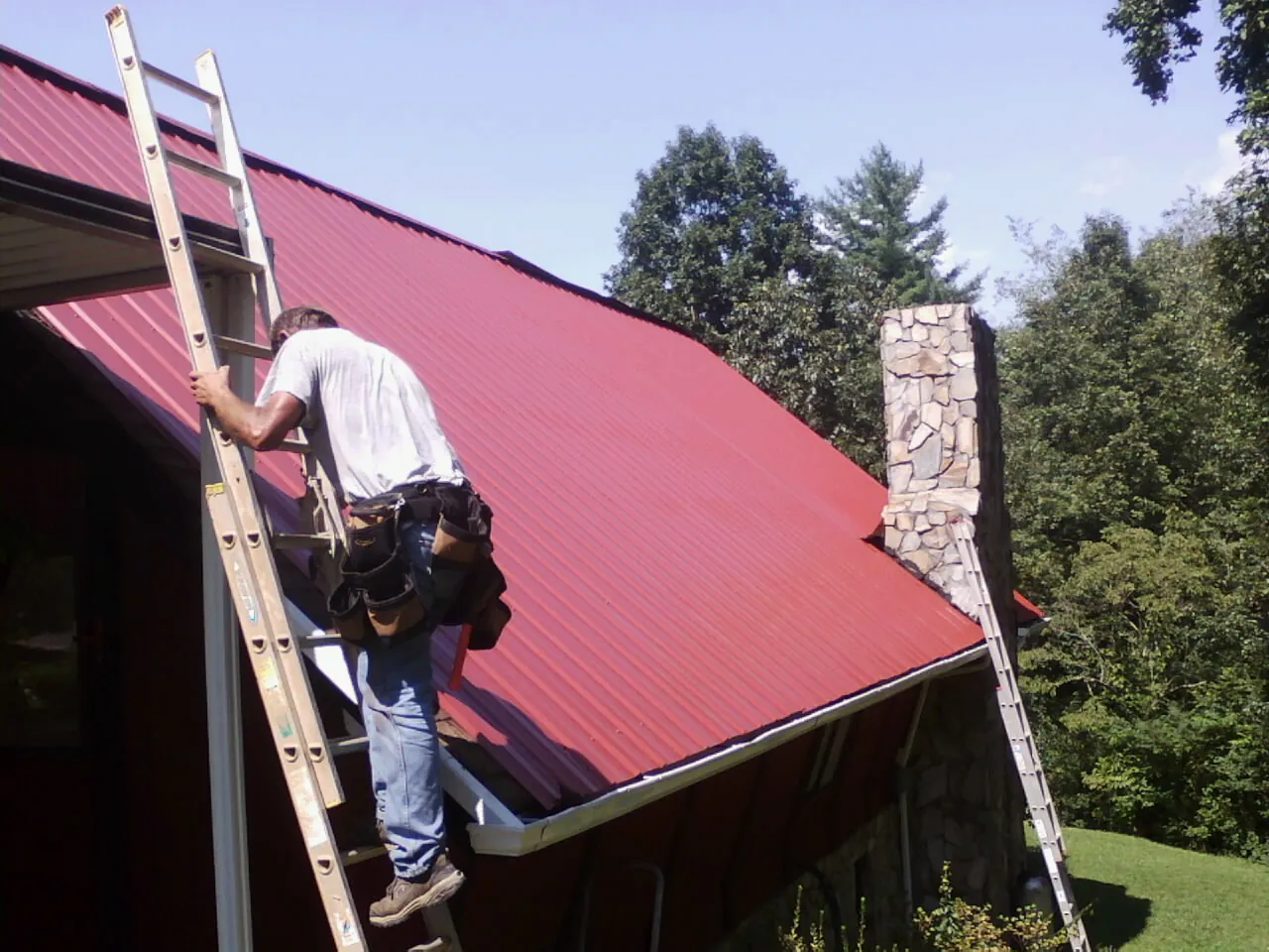 Roofer on a ladder beside a red metal roof and stone chimney — Soffit & Fascia Repair by Roofing Brothers in Reelsville